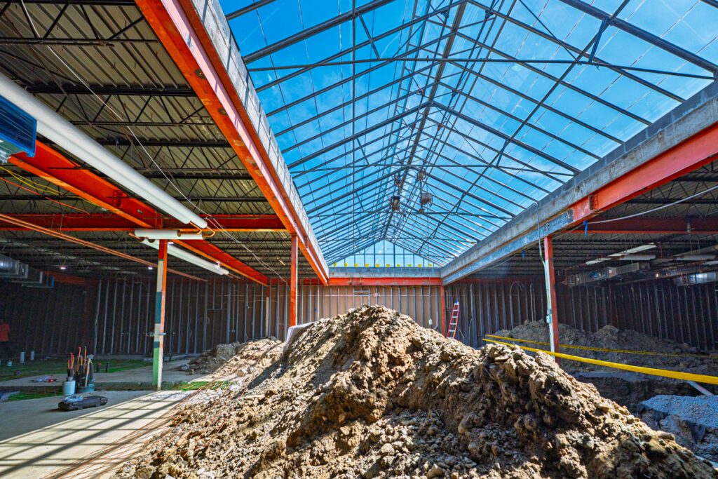 A construction site inside a large building with a glass roof, red steel beams, dirt piles, pipes, and a ladder, brightly lit by natural sunlight.
