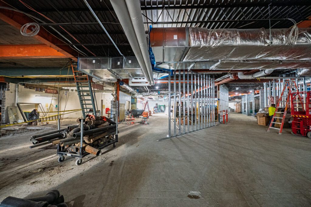 Construction site interior with exposed pipes, ducts, and metal wall frames. Ladders, construction materials, and equipment are scattered on the dusty floor under a partially finished ceiling.