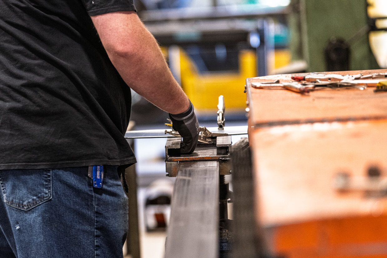 A person wearing jeans and a black shirt works with metal using a clamp on an orange workbench in a workshop, possibly referencing Building Information Modeling principles for precision. Tools are scattered on the bench, and only the person's torso and arms are visible.