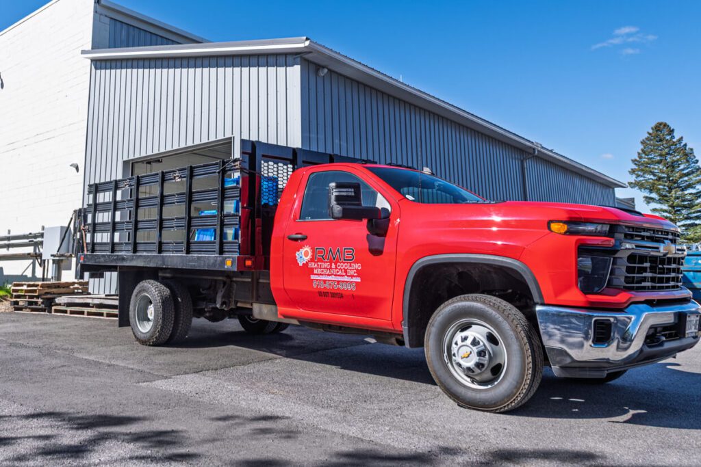 A red flatbed truck with RMS Roofing & Siding branding is parked beside a large gray industrial building on a sunny day. The truck bed has black metal siding, and trees are visible in the background—a scene that highlights the role of Building Information Modeling in modern construction projects.