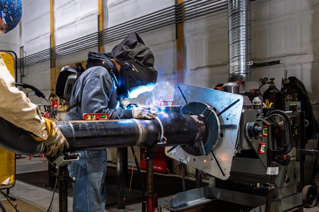 A worker wearing protective gear welds a large metal pipe in an industrial workshop, with welding sparks visible and various tools and equipment around, demonstrating how Building Information Modeling can enhance efficiency and safety in complex fabrication environments.
