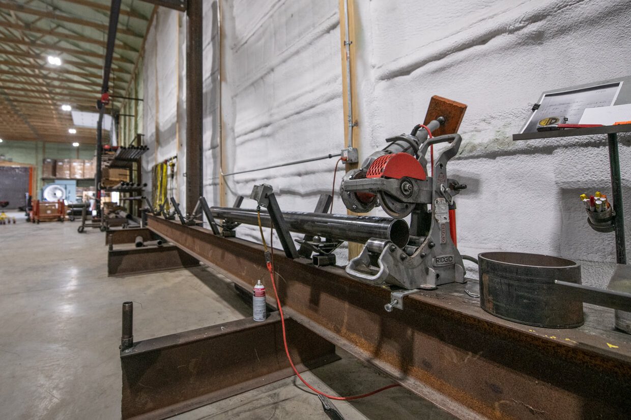 A metal-cutting saw is mounted on a long steel beam inside a spacious industrial workshop designed using Building Information Modeling. Shelving, equipment, and metal parts are visible in the background. The area is well-lit with high ceilings.