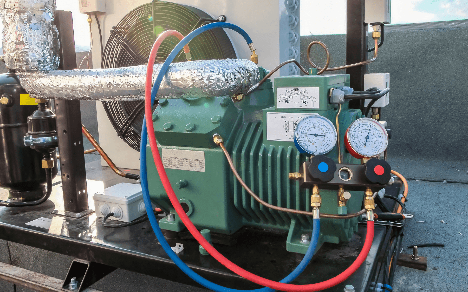 A close-up of a commercial refrigeration HVAC compressor unit on a roof, showing insulated pipes, two pressure gauges, thick red and blue hoses, and various metal components.