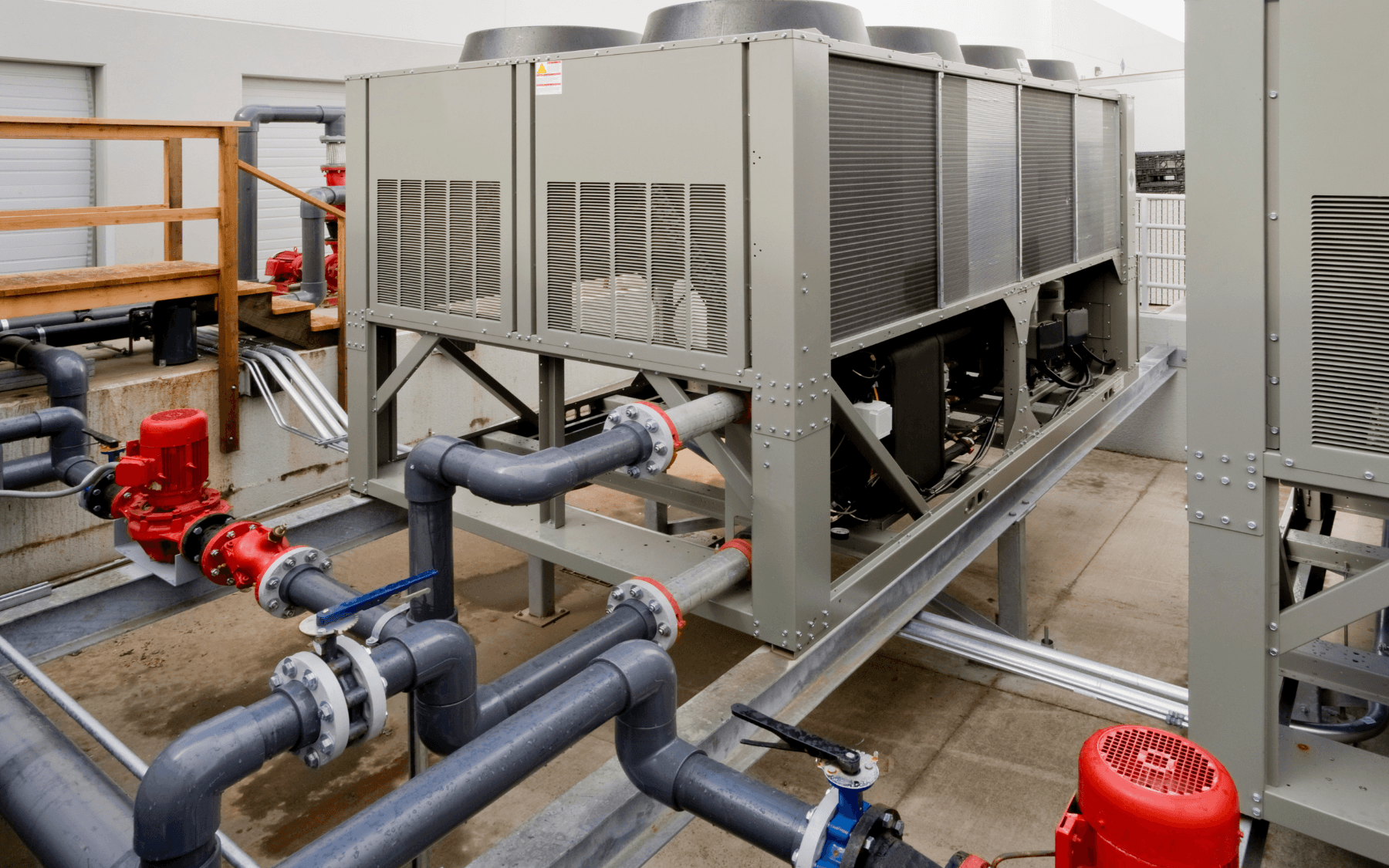 Large industrial HVAC chiller unit with metal casing, visible fans, pipes, and red valves—designed for Commercial Refrigeration—installed outdoors on a concrete pad near a building and wooden railing.
