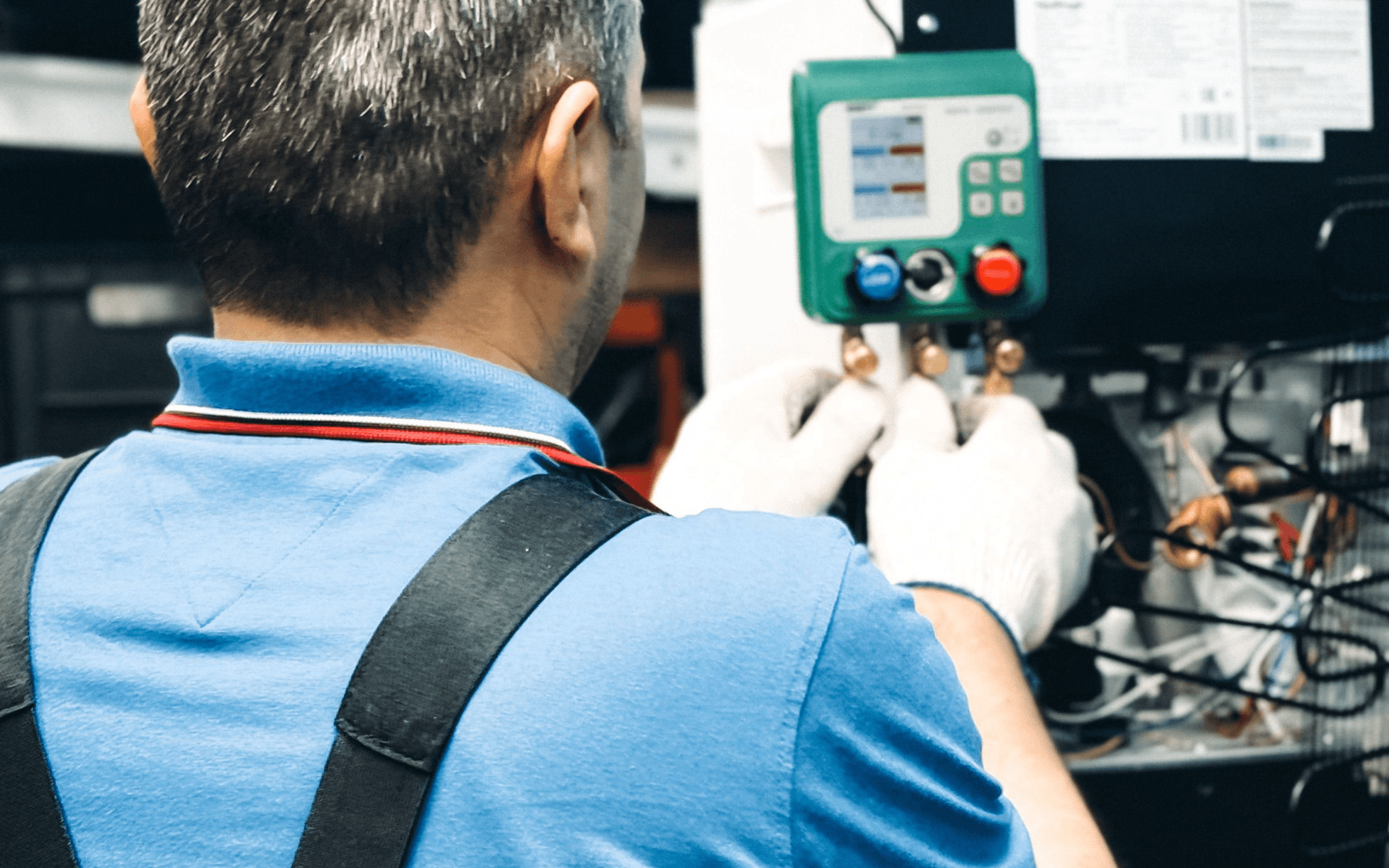 A technician wearing a blue shirt and white gloves adjusts controls on a green industrial panel or machine, working with various wires and equipment in a commercial refrigeration setting.