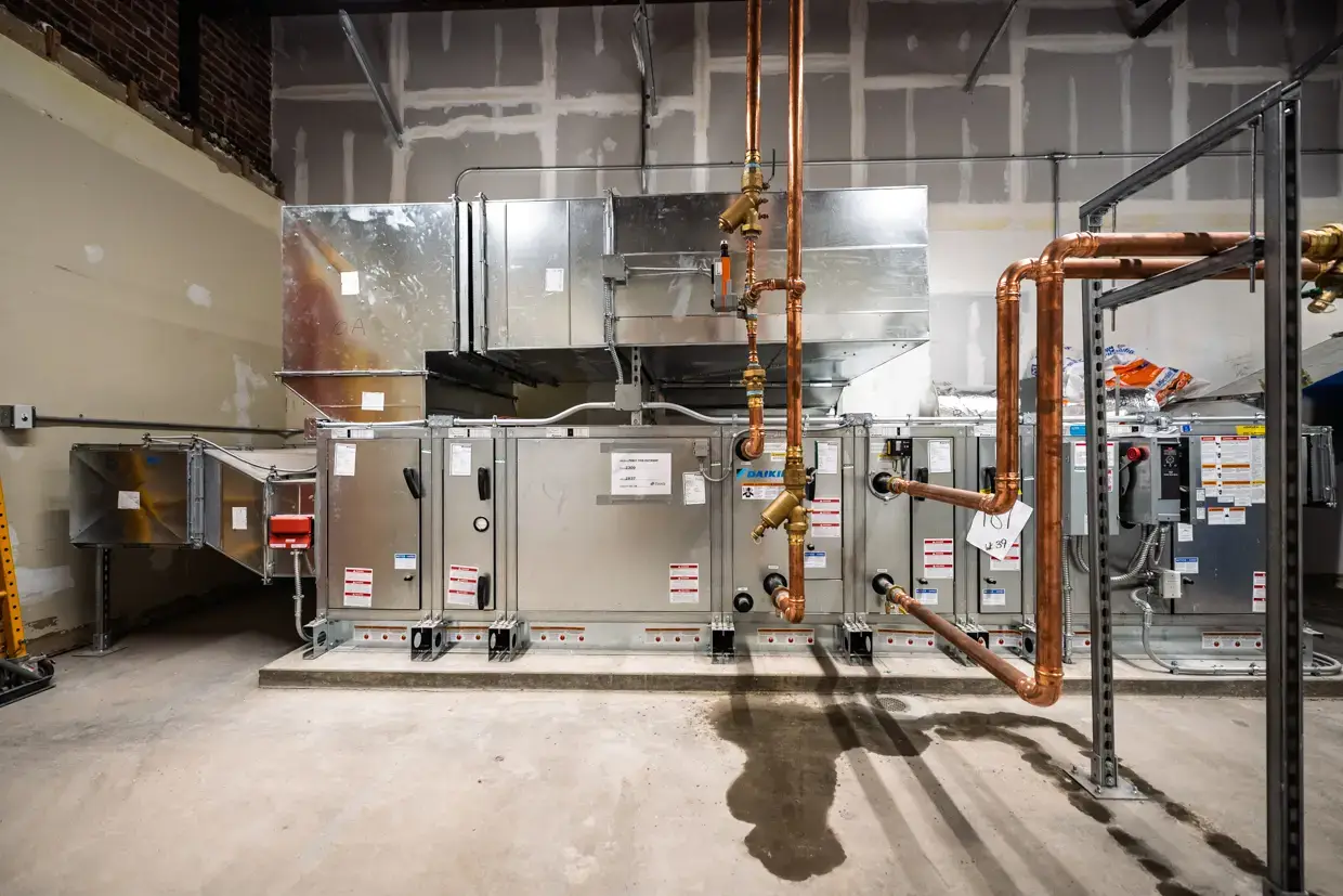 Large industrial HVAC system with silver ductwork and exposed copper pipes is installed in a room with unfinished walls and concrete floor. Labels and warning signs are visible on the equipment, highlighting the importance of preventative maintenance for reliable operation and safety.