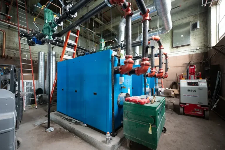 Large blue industrial boilers with red valves and black pipes in a utility room, surrounded by tools, a green cart, a ladder, and various maintenance equipment essential for preventative maintenance.