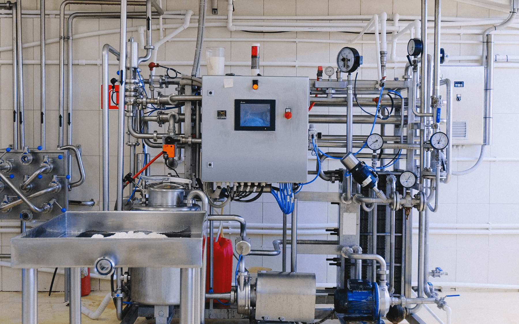 Industrial control panel with process piping, pipes, gauges, valves, and a touchscreen display in a clean, tiled room. A stainless steel sink is in the foreground, with powder or residue inside. Various hoses and wiring are visible.