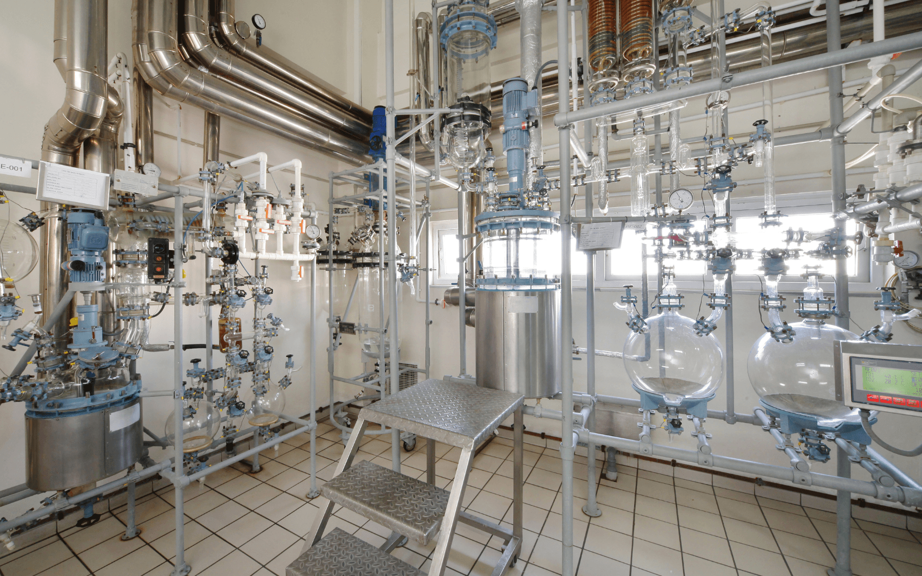 A modern laboratory with glass reactors, metal pipes, process piping, valves, gauges, and a metal staircase, used for chemical or pharmaceutical processing. The room has tiled floors and bright lighting.