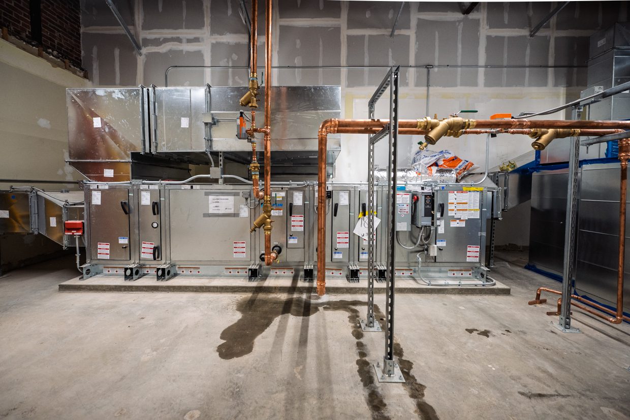 Large HVAC units are installed in a utility room with exposed pipes, ductwork, and unfinished walls. The concrete floor shows a small water leak beneath the pipes.