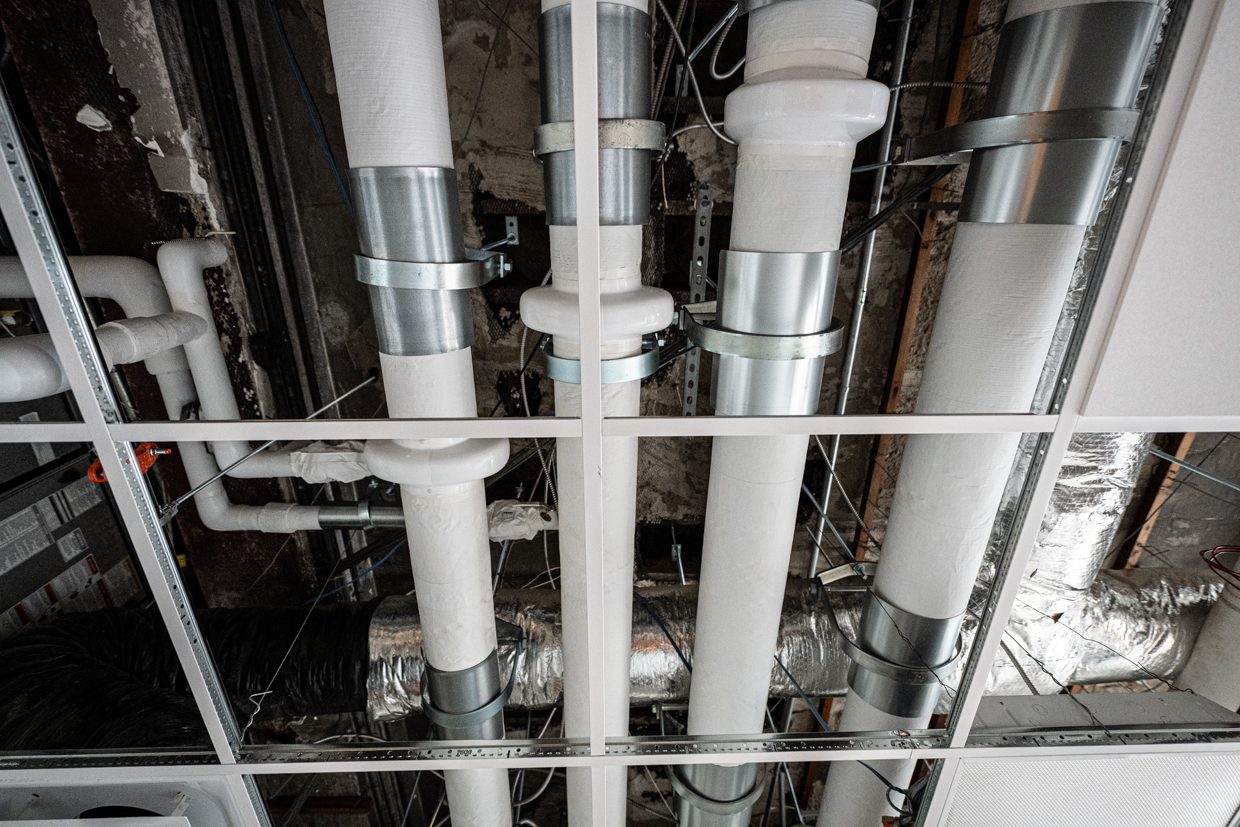 Exposed ventilation ducts and pipes run through a drop ceiling grid, showing various metal and white plastic components in an unfinished building interior.
