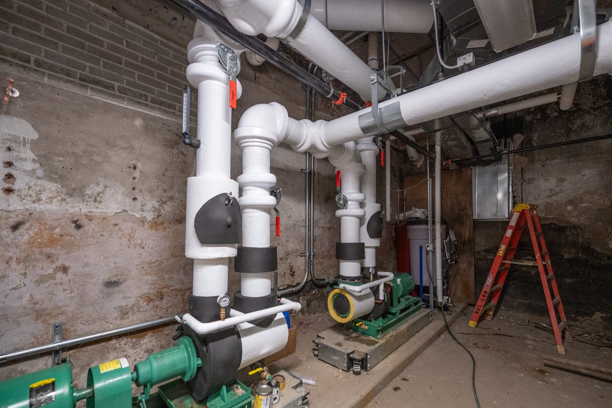 Two large insulated pipes connected to green pumps in a basement with exposed brick walls. A red ladder stands in the background near a water heater and other utility equipment.