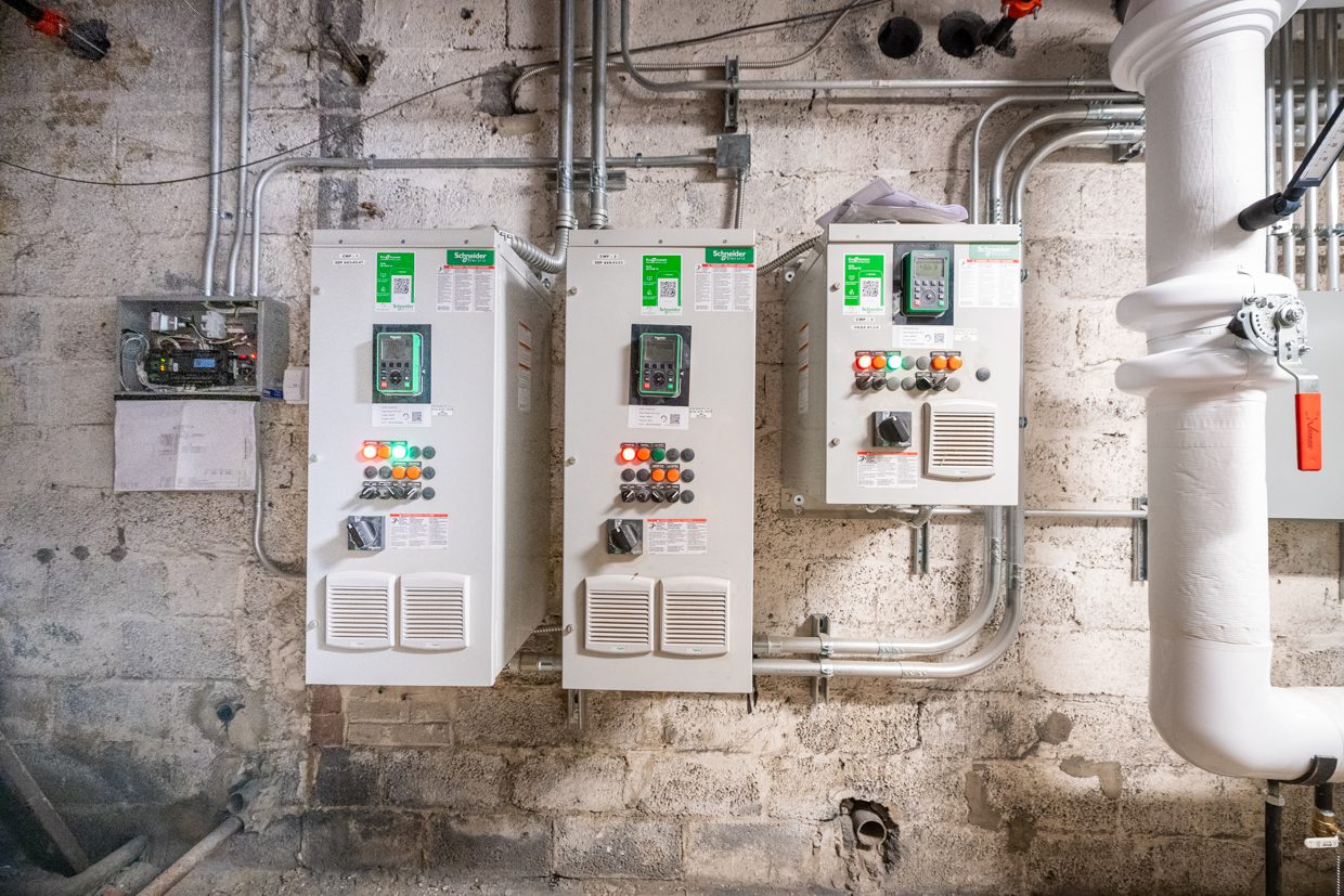 Three electrical control panels with buttons and switches are mounted on a concrete wall, surrounded by exposed pipes and wiring, in an industrial or utility room setting.