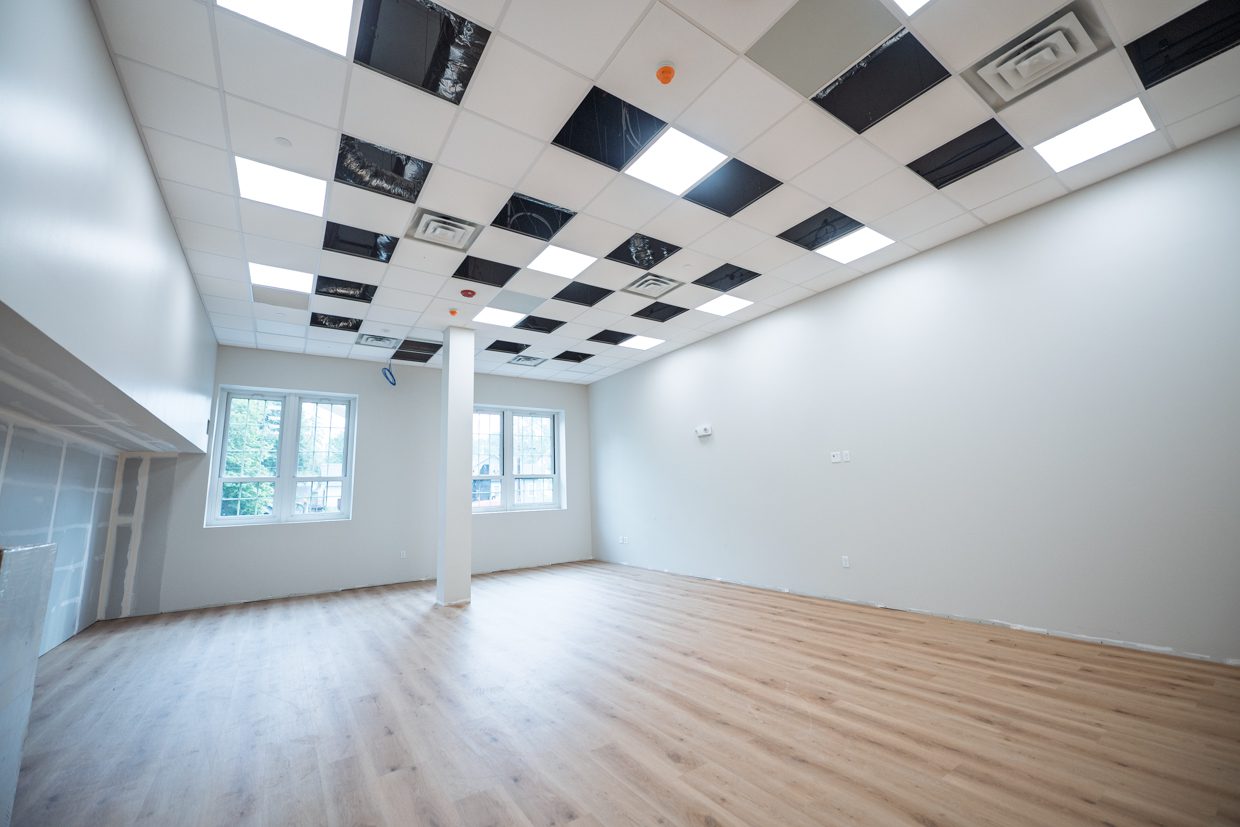 Bright, empty room with light wood flooring, white walls, large windows, and a ceiling with alternating black and white panels, some of which are missing, exposing wires and ductwork.
