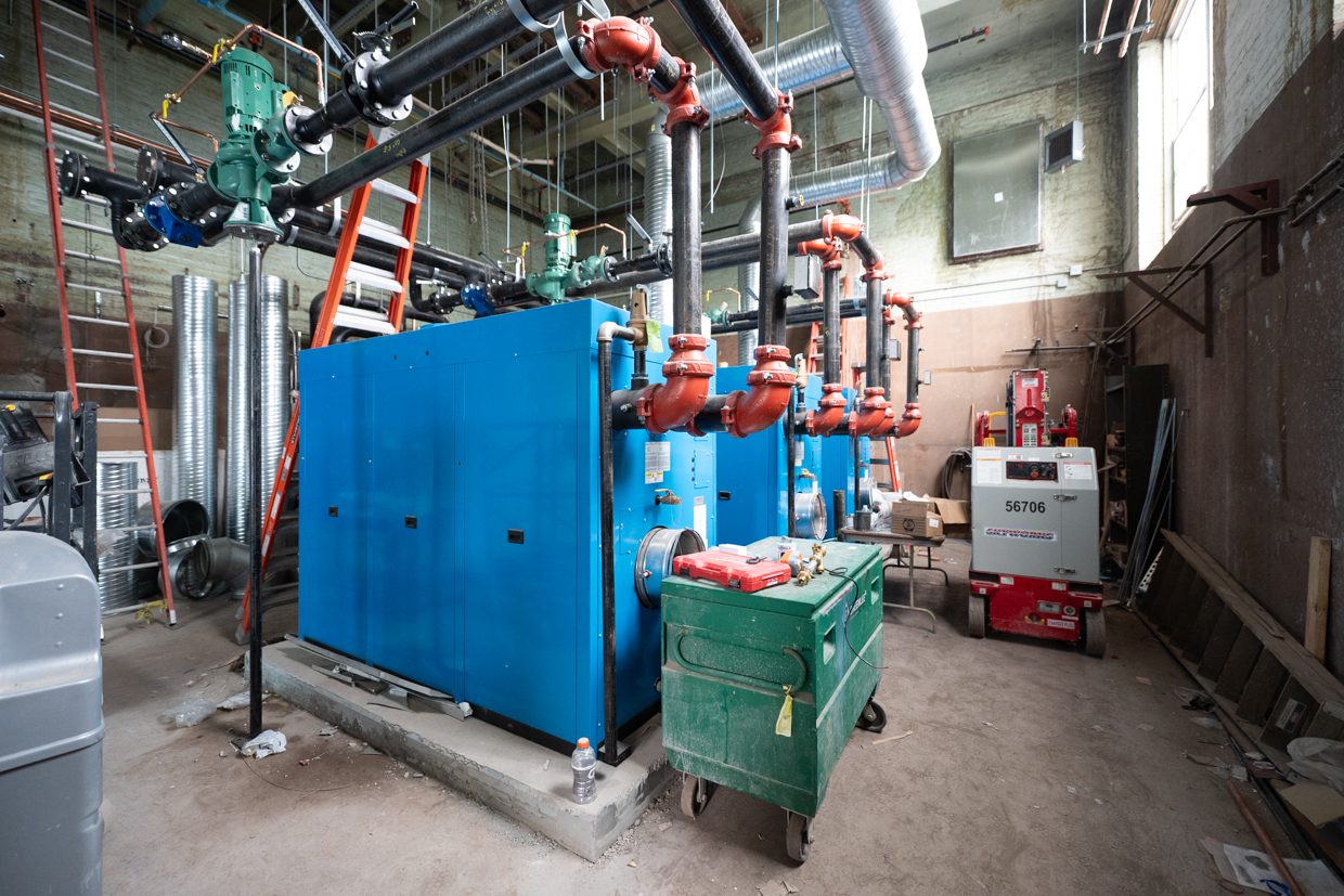 Large blue industrial machines with pipes and valves in a mechanical room under construction, surrounded by tools, ladders, and construction equipment on a dusty concrete floor.