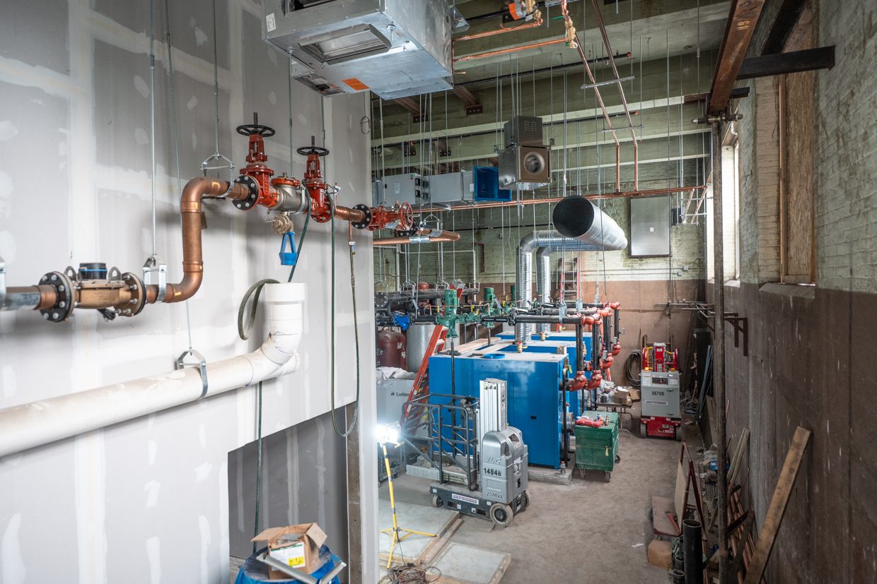 Industrial room with exposed pipes, blue machinery, and ventilation ducts. Construction materials, ladders, and tools are scattered around. The space appears unfinished with visible insulation and partially painted walls.