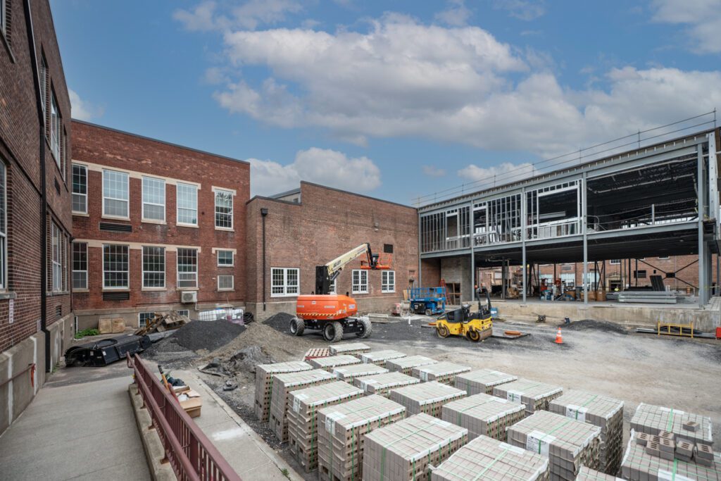 A construction site shows an addition being built onto a brick building, with scaffolding, construction vehicles, and stacks of bricks in the yard under a partly cloudy sky.