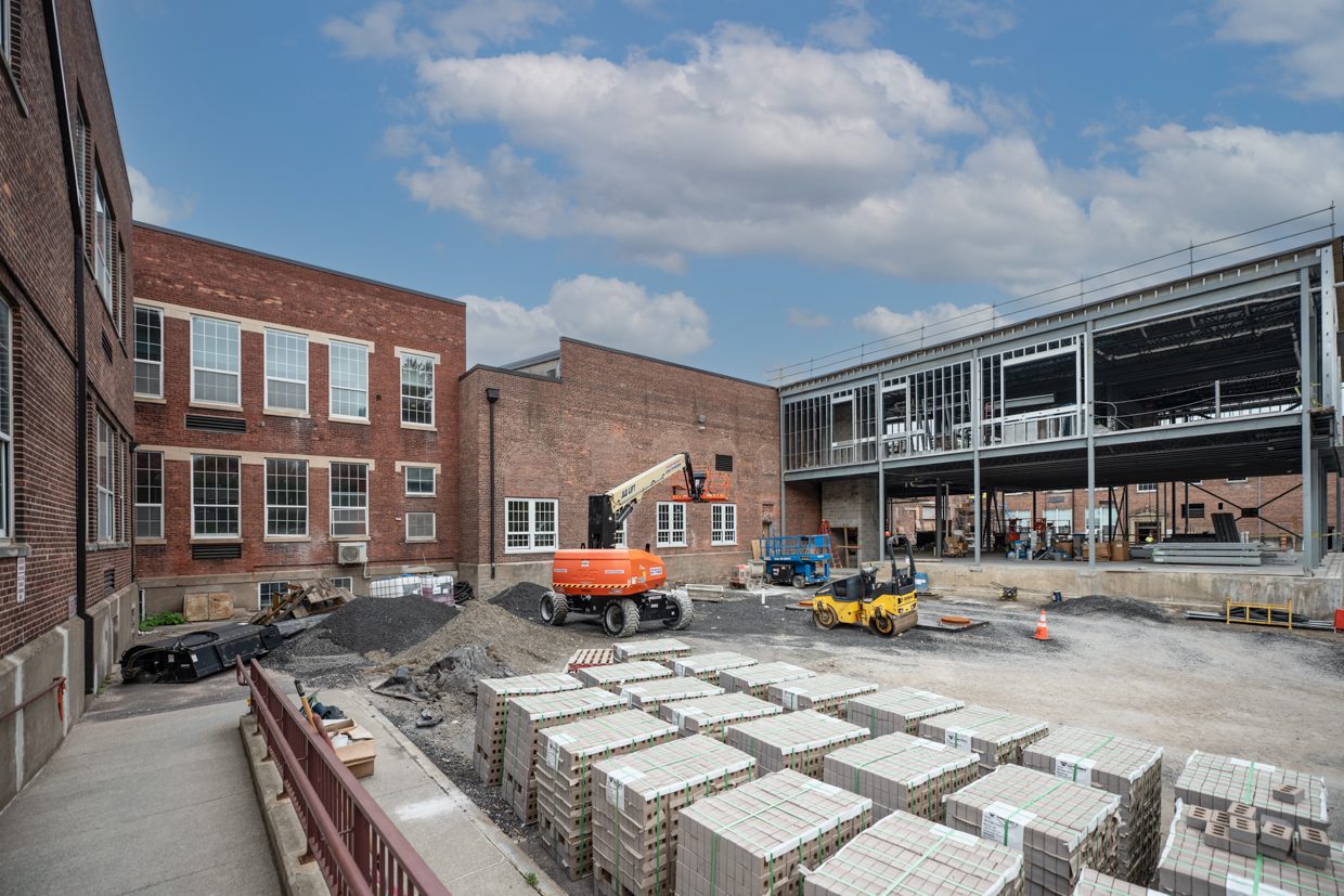 A construction site shows an addition being built onto a brick building, with scaffolding, construction vehicles, and stacks of bricks in the yard under a partly cloudy sky.