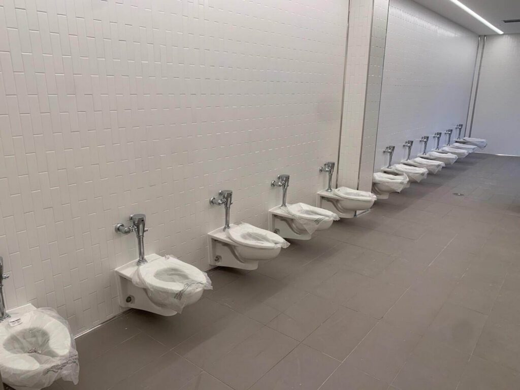A row of wall-mounted toilets with plastic covers lines a tiled, white-walled restroom. Featuring an open layout with no stalls or partitions, the space highlights a streamlined approach to commercial plumbing.