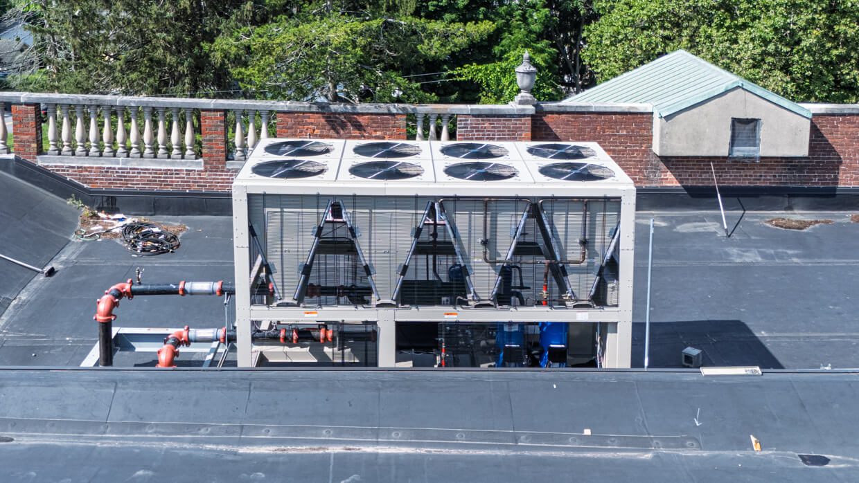 Large HVAC unit with multiple fans on a flat rooftop, surrounded by pipes and a brick wall—part of a comprehensive design build project—with trees and a building featuring a green roof in the background.