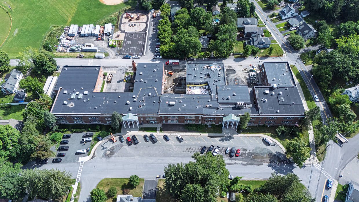 Aerial view of a large school building, featuring a Design Build layout with a parking lot in front, basketball court and playground in back, all surrounded by trees, houses, and roads.