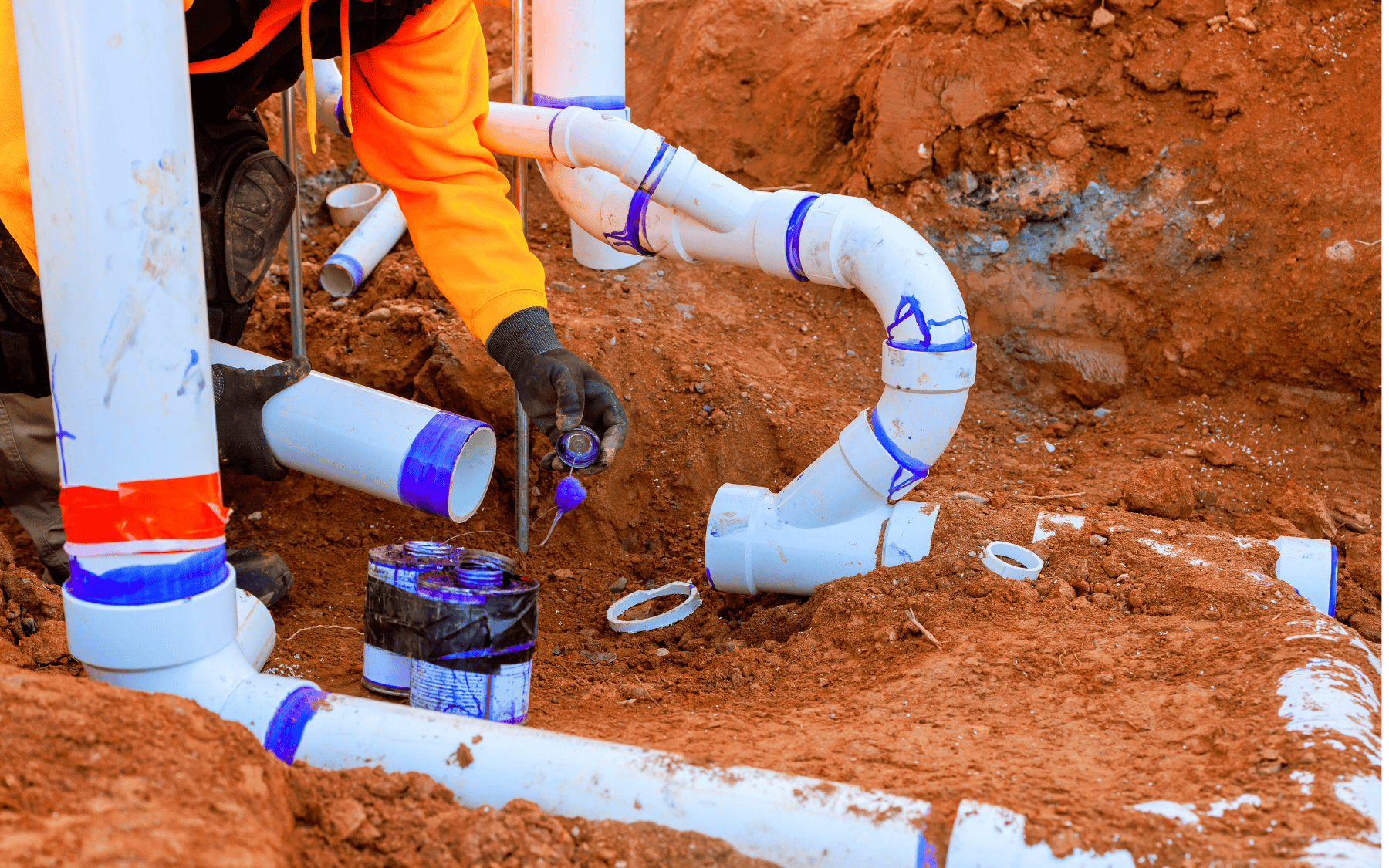 A person in an orange jacket applies glue to PVC pipes while installing commercial plumbing in a trench filled with reddish-brown soil.