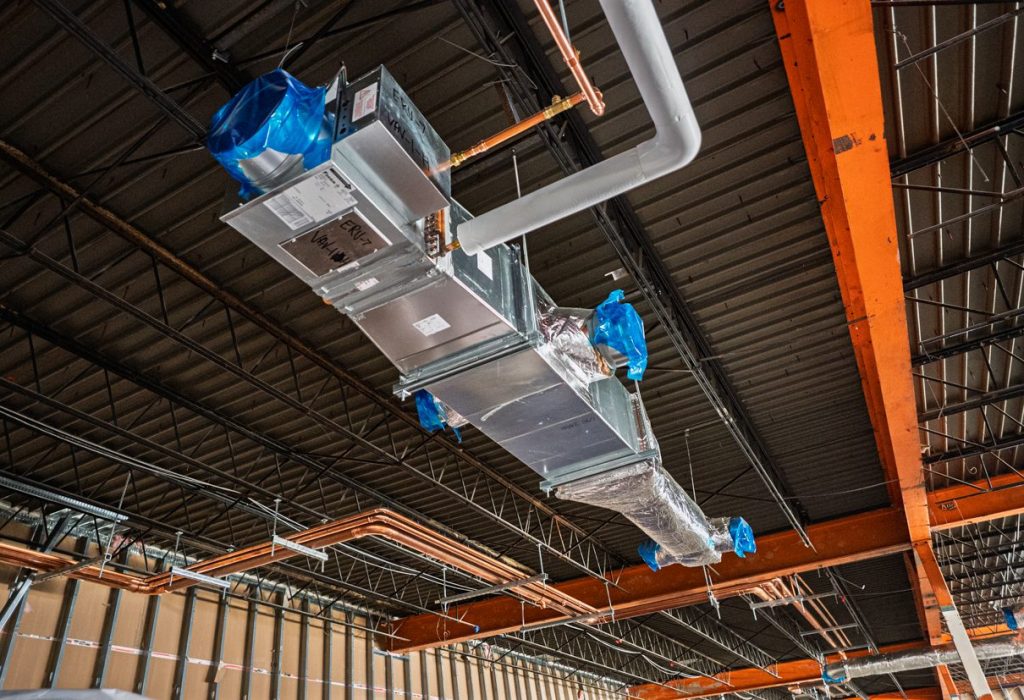 Exposed Mechanical Service HVAC ductwork and pipes with blue plastic coverings hang from the ceiling of a building under construction, with visible beams and framing.
