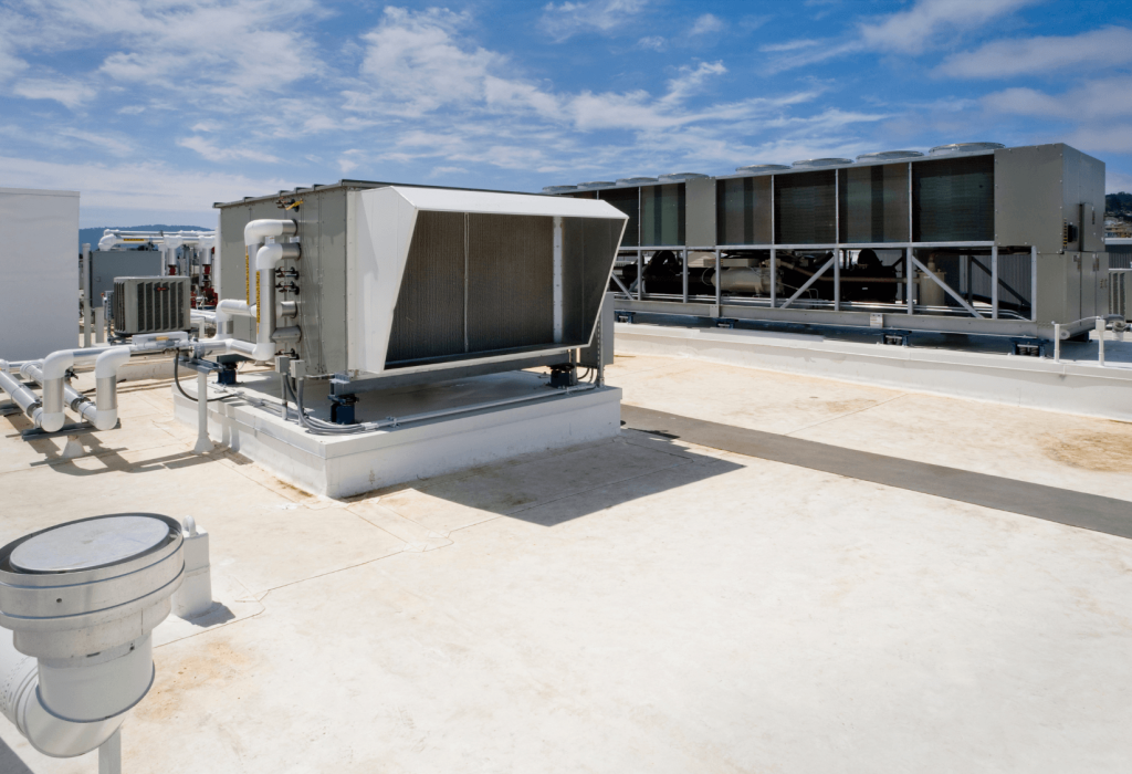 Large HVAC units and ventilation systems, part of a comprehensive mechanical service installation, are arranged on a flat commercial rooftop under a partly cloudy sky. Distant landscape and buildings are visible in the background.