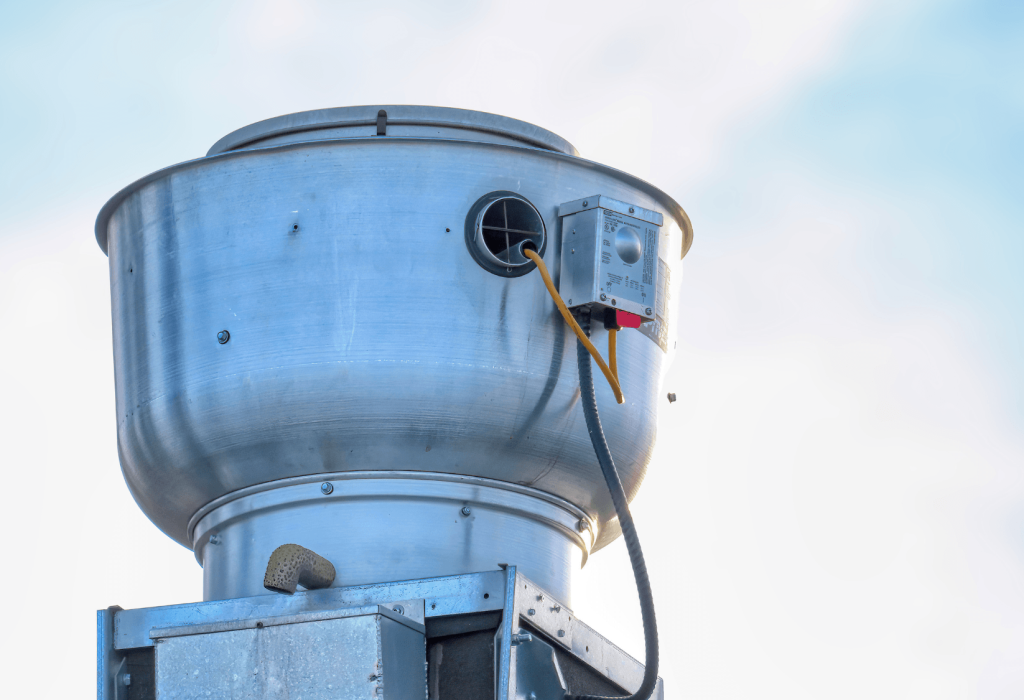 A close-up view of a large industrial exhaust fan or ventilation unit mounted on a rooftop, with visible electrical wiring and a partly cloudy sky in the background, highlighting essential components of mechanical service.