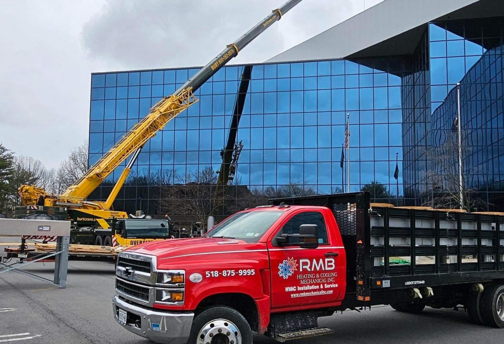 A red RMB Restoration & Cleaning truck is parked in front of a modern glass building, while a yellow crane lifts HVAC materials in the background on a cloudy day.