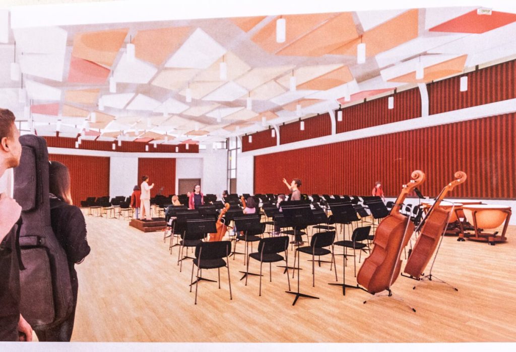 Musicians gather in a bright rehearsal room with red acoustic panels, geometric ceiling shapes, and empty black chairs surrounded by various instruments—including cellos and timpani—while an efficient HVAC system maintains a comfortable atmosphere as the conductor stands on the podium.