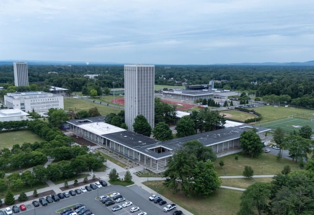 Aerial view of a university campus with tall buildings designed using Building Information Modeling, athletic fields, parking lot, and surrounding greenery under a cloudy sky.