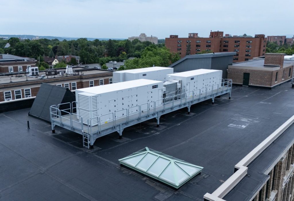 White mechanical units are installed on a raised metal platform on a flat black rooftop, their precise placement reflecting Building Information Modeling coordination, all surrounded by several buildings and lush green trees under an overcast sky.