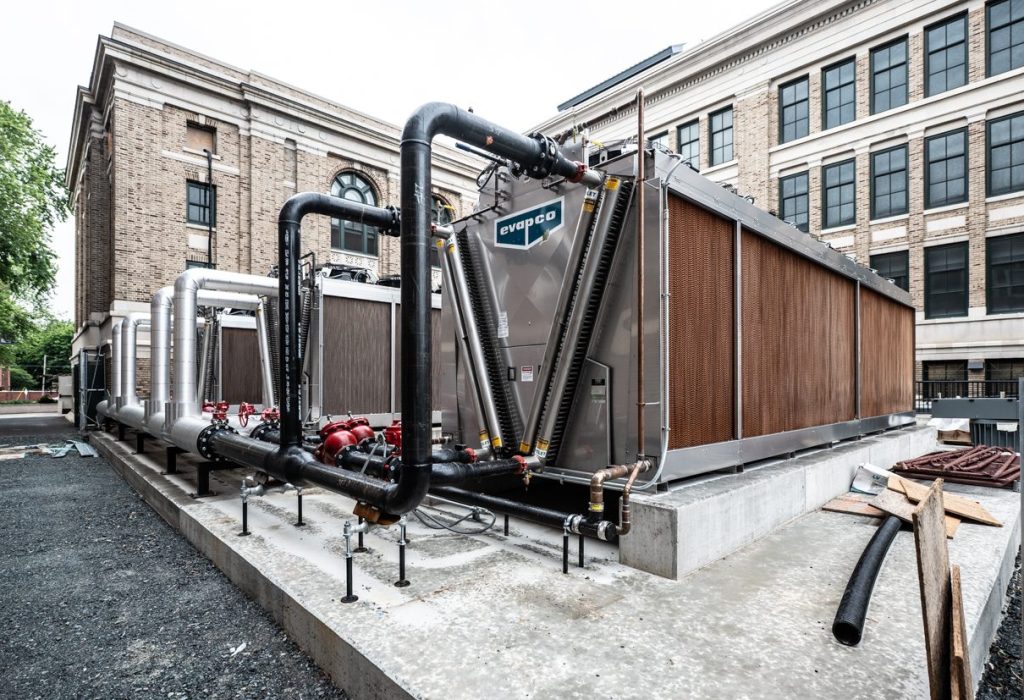 Large industrial HVAC cooling units with exposed pipes and pumps are installed on a concrete platform outside a multi-story brick building with large windows. Construction materials and gravel are visible nearby, highlighting a project that likely benefited from Building Information Modeling to coordinate such complex mechanical systems and site logistics.