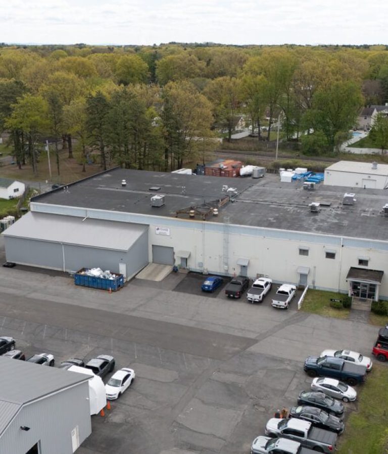 Aerial view of a large industrial building with a parking lot, several parked cars, and trees in the background, showcasing how Building Information Modeling can optimize the layout of shipping containers and equipment near the building.