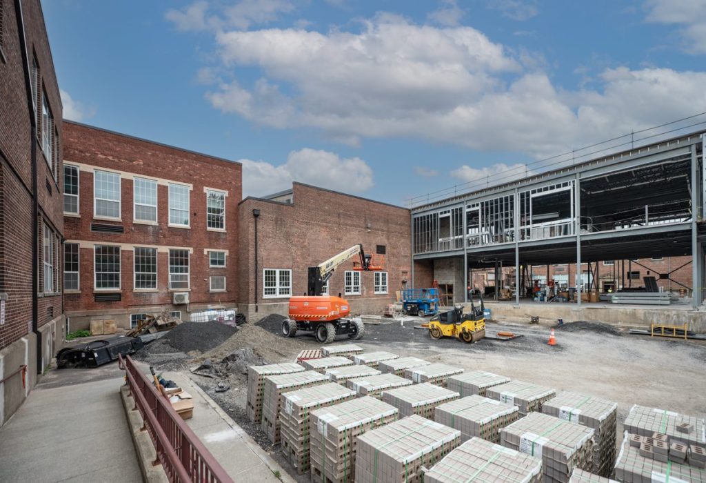 A construction site shows an addition being built onto a brick building, with scaffolding, construction vehicles, and stacks of bricks in the yard under a partly cloudy sky.